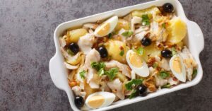 Prato de bacalhau gomes da costa com batatas, pimentões e azeitonas, decorado com ovos cozidos em uma travessa.
