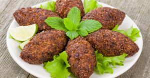 Prato com kibbeh ou quibes fritos servidos sobre folhas de alface, decorados com folhas de hortelã e fatias de limão ao lado