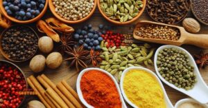 Variedade de temperos e especiarias dispostos sobre uma mesa de madeira, incluindo canela em pau, pimenta em grãos, cardamomo, anis-estrelado, noz-moscada, cúrcuma em pó e páprica.