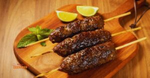 Espetinhos de kafta grelhados sobre uma tábua de madeira, decorados com coentro fresco e acompanhados por pequenos potes de molhos ao fundo.