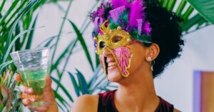 Mulher usando uma máscara colorida e segurando um drink de Carnaval, em um ambiente festivo com decoração tropical.