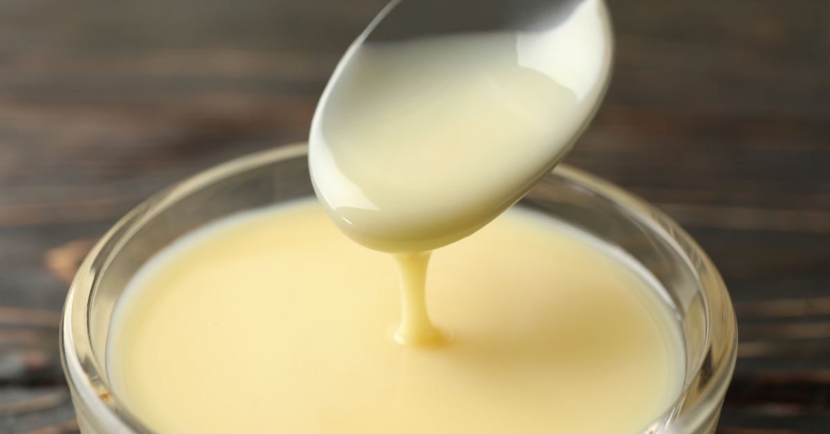 Colher de leite condensado sendo derramado em um recipiente de vidro sobre uma mesa de madeira escura.