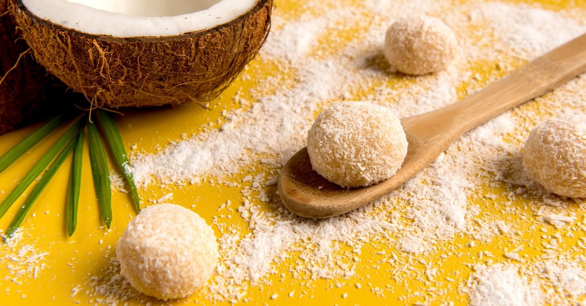 Beijinho de coco com coco ralado em superfície amarela, acompanhados de meio coco e folhas verdes ao fundo.