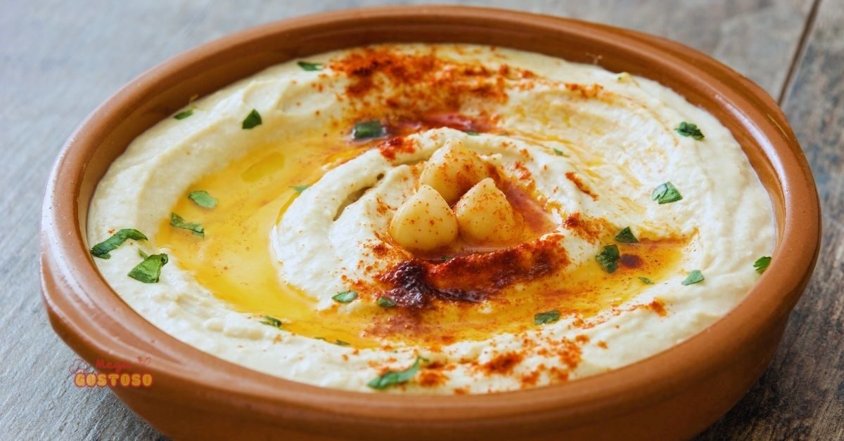 Prato de hummus servido em tigela de cerâmica, decorado com azeite, páprica, grãos de grão-de-bico e folhas de salsa.